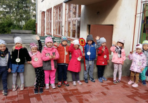 Dzieci z gruy drugiej o nazwie ,,Biedroneczki" stoją na tle budynku przedszkola i prezentują swój emblemat.