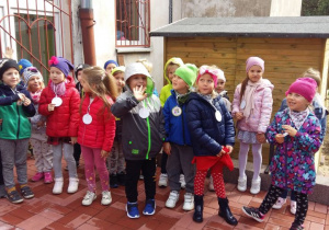 Dzieci z grupy czwartej o nazwie ,,Mróweczki" stoją na tle budynku przedszkola i prezentują swój emblemat.