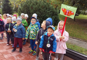 Dzieci z grupy trzeciej o nazwie ,,Motylki" stoją na tle ogrodu przedszkolnego i prezentują swój emblemat.