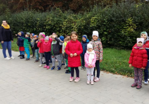 Przedszkolaki stoją w grupie wraz z opiekunem na tle zieleni.