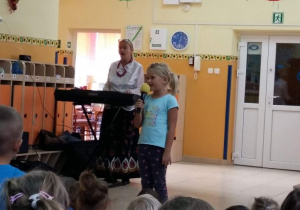Dziewczynka z grupy piątej stoi z mikrofonem i śpiewa piosenkę dla pozostałych przedszkolaków.