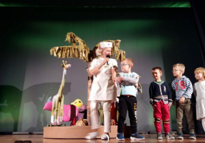 Czworo dzieci stoi na scenie wraz z aktorką w stroju pielęgniarki. Dzieci biorą udział w konkursie i odpowiadają na pytania zadawane przez aktorkę.