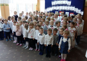 Przedszkolaki ubrane w odświętne stroje, stoją na tle dekoracji z napisem „Święto Niepodległości” i śpiewają hymn Polski.