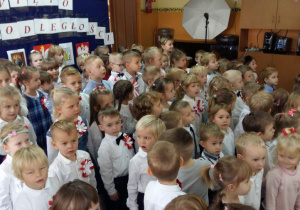 Przedszkolaki ubrane w odświętne stroje, stoją na tle dekoracji z napisem „Święto Niepodległości” i śpiewają hymn Polski.