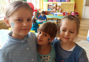 3 dziewczynki z niebieskimi sercami na twarzy stoją blisko siebie w sali przedszkolnej. .