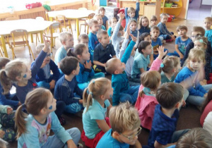 Dzieci ubrane w stroje w kolorze niebieskim, siedzą na dywanie przed tablicą interaktywną. Odpowiadają na pytania nauczyciela na temat praw dziecka.