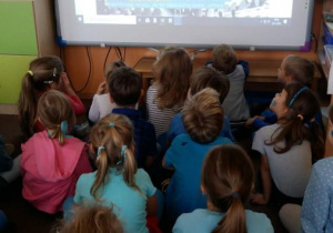 Dzieci ubrane w stroje w kolorze niebieskim, siedzą na dywanie przed tablicą interaktywną. Oglądają prezentację o prawach dziecka