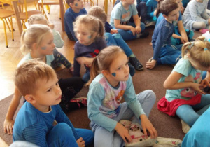 Dzieci ubrane w stroje w kolorze niebieskim, siedzą na dywanie przed tablicą interaktywną. Oglądają prezentację o prawach dziecka.