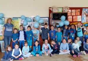 Dzieci z grupy 5 ubrane w stroje w kolorze niebieskim oraz nauczycielka, pozują do zdjęcia na tle dekoracji, przedstawiającej plakaty z prawami dziecka w sali przedszkolnej.