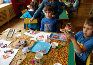 Dzieci siedzą przy stoliku przedszkolnym. Wykonują prace plastyczne przedstawiające dzieci z innych krajów w strojach ludowych, tradycyjnych.