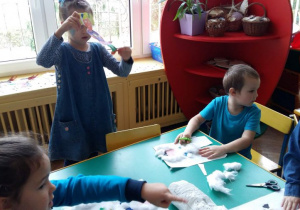 Dzieci siedzą przy stoliku przedszkolnym. Wykonują prace plastyczne przedstawiające dzieci z innych krajów w strojach ludowych, tradycyjnych. W tle widać kącik przyrody.