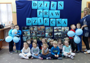 Dzieci z grupy 2 ubrane w stroje w kolorze niebieskim, pozują do zdjęcia na tle dekoracji, przedstawiającej plakaty z prawami dziecka w szatni przedszkolnej. Czworo dzieci w rękach trzyma błękitne balony.
