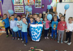 Dzieci z grupy IV stoją na tle tablicy interaktywnej z wykonana praca plastyczną. Kilkoro dzieci trzyma w rekach błekitne balony.