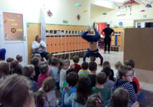 Tancerz przedstawia taniec Breakdance. Dzieci siedzą na dywanie i przyglądają się występom artystów.W tle widoczne są szafki przedszkolne w szatni.