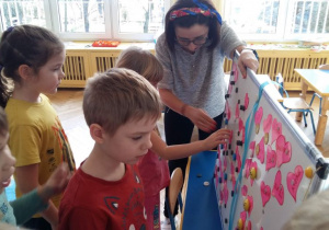 Grupka dzieci i nauczycielka stoją przed białą tablicą. Nauczycielka trzyma tablicę magnetyczną, na której mieszczą się serduszka z imionami. Chłopiec ubrany w pomarańczową bluzkę losuje 1 karteczkę. Na kartce widnieje imię dziewczynki.