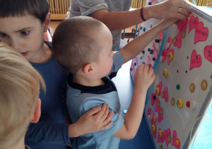 Dzieci stoją przed białą tablicą magnetyczną, na której mieszczą się serduszka z imionami. Chłopiec wybiera 1 serduszko z wróżbą.