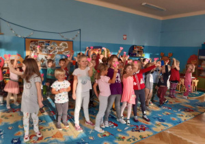 Dzieci z dwóch grup (trzeciej i piątej) uczestniczą w zabawie andrzejkowej. Dzieci pokazują wylosowane w postaci serc wróżby andrzejkowe. W tle widoczne są meble przedszkolne i zabawki.