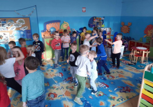 Dzieci z dwóch grup (trzeciej i piątej) uczestniczą w zabawie andrzejkowej. Tańczą w parach lub rozsypce w sali przedszkolnej. W tle widoczne są meble przedszkolne i zabawki.