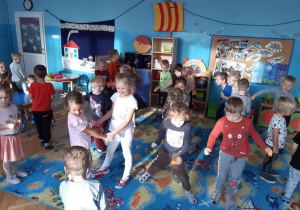 Dzieci z dwóch grup (trzeciej i piątej) uczestniczą w zabawie andrzejkowej. Tańczą w parach lub rozsypce w sali przedszkolnej. W tle widoczne są meble przedszkolne i zabawki.