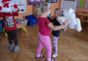 Dzieci tańczą z pluszowymi misiami. W tle widoczne są tablice dydaktyczne, meble przedszkolne i zabawki.