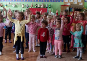 Przedszkolaki stoją na środku sali, ćwiczą układ taneczny, prezentowany przez baletnicę. Mają uniesione do góry ręce.