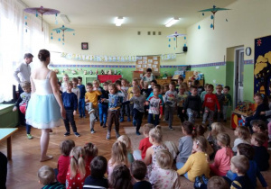 Chłopcy stoją na środku sali, ćwiczą układ taneczny, prezentowany przez baletnicę.