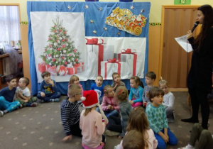 Dzieci siedzą na dywanie. Pani prowadząca zabawę mikołajkową trzyma w ręku mikrofon. W tle dekoracja świąteczna.