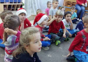 Dzieci siedzą na dywanie. Niektóre z nich mają na głowie czapkę Mikołaja.