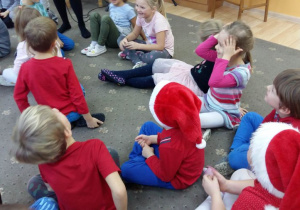 Dzieci siedzą na dywanie podczas zabawy mikołajkowej. Niektóre z nich mają na głowie czapkę Mikołaja.