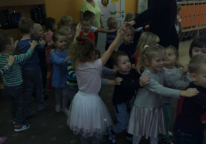 Dzieci bawią się podczas zabawy mikołajkowej. Dzieci poruszają się po sali ustawione jedno za drugim w tzw. „pociąg”. Prowdząca bal wspólnie z przedszkolakiem "tworzą most" pod którym przechodzą dzieci..