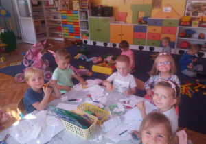 Grupa przedszkolaków siedzi przy stole. Na stoliku rozłożone są papiery do wycinania, nożyczki, szablony do odrysowywania, mazaki, kredki. W tle widać dzieci bawiące się na dywanie.
