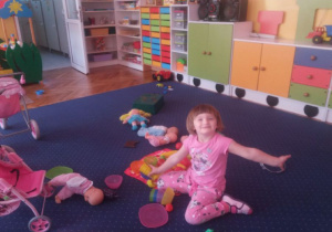 Dziewczynka bawi się zabawkami na dywanie. W tle widoczne są meble przedszkolne i zabawki