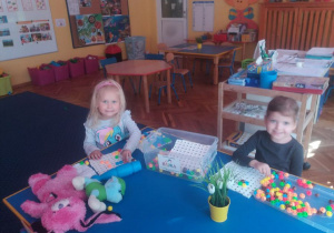 2 dziewczynki bawią się klockami przy stoliku. W tle widoczne są meble przedszkolne i zabawki