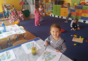 Dziewczynka maluje farbami. W tle widać dzieci bawiące się na dywanie.