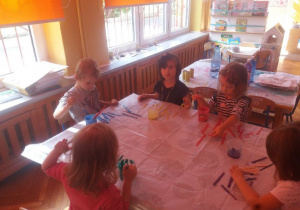 Grupa przedszkolaków siedzi przy stole. Na stoliku rozłożone są patyczki drewniane i farby. .