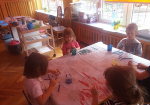 Grupa przedszkolaków siedzi przy stole. Na stoliku rozłożone są patyczki drewniane i farby.