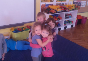 Czworo dzieci przytula się do siebie. W tle widoczne są meble przedszkolne i zabawki i tablica interaktywna.
