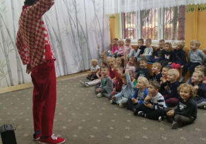 Dzieci siedzą na dywanie. Przed nimi na scenie w szatni przedszkolnej stoi komik przebrany w kraciastą koszulę , czerwone spodnie i czarny kapelusz. Prezentuje program artystyczny dla dzieci w przedszkolu.
