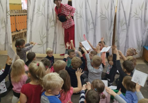 Dzieci siedzą na dywanie z podniesionymi do góry kartkami. Przed nimi na scenie w szatni przedszkolnej stoi komik przebrany w kraciastą koszulę , czerwone spodnie i czarny kapelusz. Prezentuje program artystyczny dla dzieci w przedszkolu.