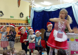 Aktorka prezentuje prosty układ tańca dzieciom stojącym na scenie. Dzieci z uwagą próbują ją naśladować.
