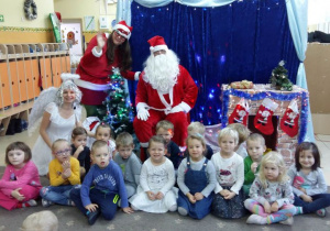 Dzieci z grupy II pozują do zdjęcia z aktorami. Mikołaj, pomocnica Mikołaja i Śnieżynka siedzą pośród dzieci. Dzieci są uśmiechnięte. W tle widoczne sa dekoracje świąteczne.