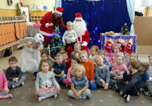 Dzieci z grupy III pozują do zdjęcia z aktorami. Mikołaj, pomocnica Mikołaja i Śnieżynka siedzą pośród dzieci. Dzieci są uśmiechnięte. W tle widoczne sa dekoracje świąteczne.