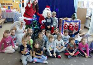 Dzieci z grupy IV pozują do zdjęcia z aktorami. Mikołaj, pomocnica Mikołaja i Śnieżynka siedzą pośród dzieci. Dzieci są uśmiechnięte. W tle widoczne sa dekoracje świąteczne.
