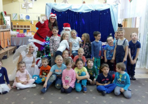 Dzieci z grupy V pozują do zdjęcia z aktorami. Mikołaj, pomocnica Mikołaja i Śnieżynka siedzą pośród dzieci. Dzieci są uśmiechnięte. W tle widoczne sa dekoracje świąteczne.