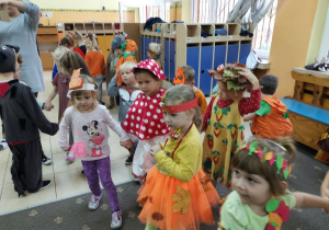 Dzieci tańczą w parach w szatni przedszkolnej na balu jesieni. Dzieci poprzebierane są w stroje muchomorków, wiewiórek, grzybków itd. W tle widoczne są szafki z ubraniami dzieci.