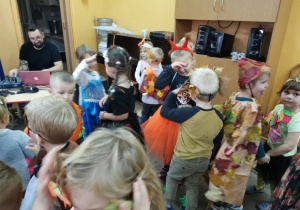 Dzieci tańczą w parach w szatni przedszkolnej na balu jesieni. Dzieci poprzebierane są w stroje lisków, wiewiórek, grzybków itd. W tle widoczne są szafki z ubraniami dzieci.