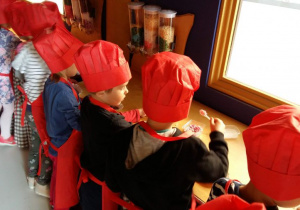 Przedszkolaki ubrane w czerwone fartuchy i czapki kucharskie dekoruje wyprodukowane przez siebie tabliczki czekolady.