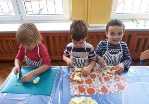 Przedszkolaki ubrane w fartuchy kuchenne siedzą przy stole i kroją na plastikowych podkładkach jabłka.