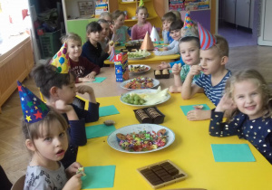 Przedszkolaki siedza przy stole. Niektóre dzieci maja na głowach urodzinowe czapeczki. Na stole ustawione sa talerzyki ze słodkościami.