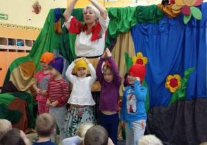 Aktorka i pięcioro przedszkolaków występuja wspólnie na scenie. Dzieci mają na głowach kolorowe czapki. W tle zawieszony jest kolorowy materiał z naszytymi żółtymi kwiatami.